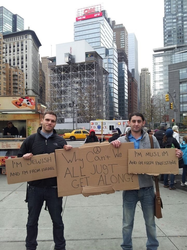 two men standing: the man on the left is holding a sign "I'M JEWISH AND I'M FROM ISRAEL" and the man on the left is holding up a sign "I'M MUSLIM AND I'M FROM PALESTINE" and they are both holding up a large sign in the middle with a peace symbol on top and the words "Why Can't We ALL JUST GET ALONG"