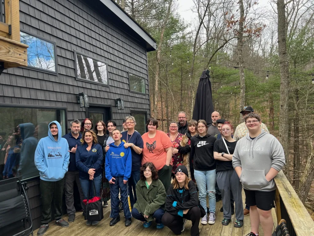 group of high school students standing on the deck of a tiny home with several staff in the back and trees in the background